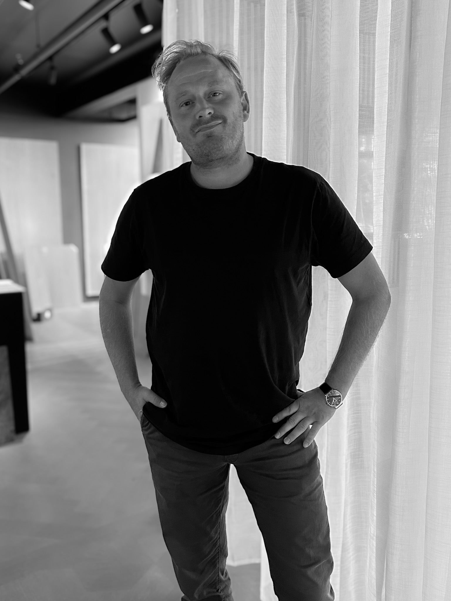 Man wearing a black t-shirt and pants standing indoors with white curtains in the background
